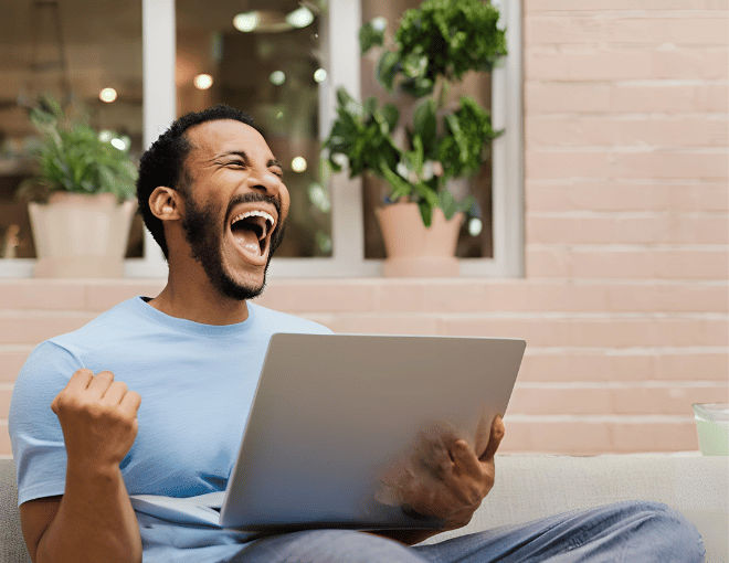 Excitedly, a man clutches his laptop, mouth wide in a triumphant grin, his posture animated with victory as he celebrates online success.